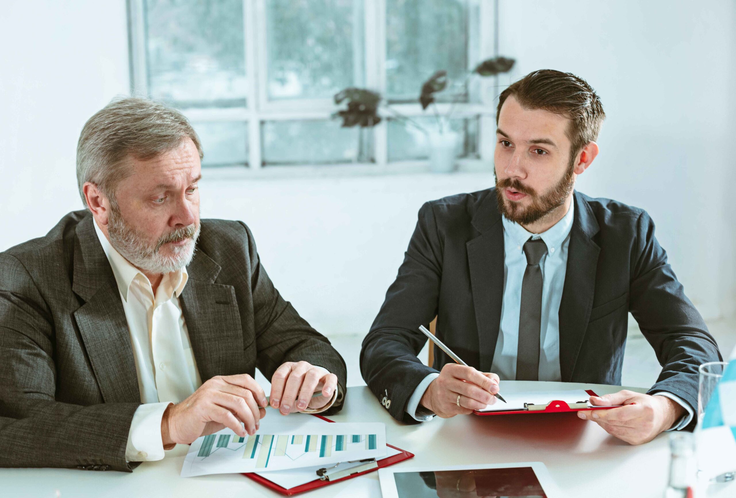 Two men in business attire discuss documents during a meeting, with charts and notes visible on the table.